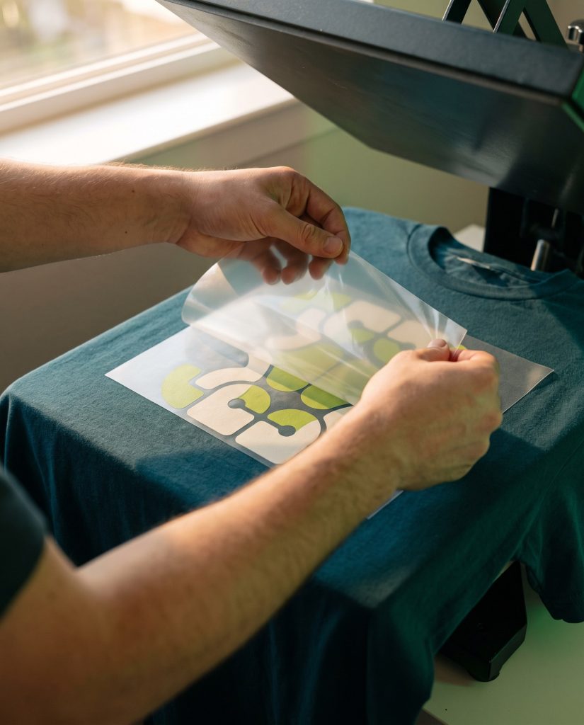 Hands peeling a DTF transfer film off a teal t-shirt on a heat press